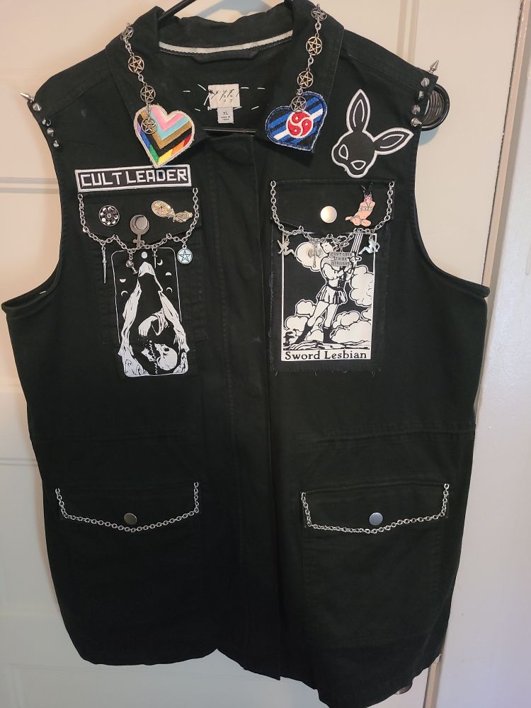 A monochrome battlevest with patches and chains. Some patches include: "Sword Lesbian" page of Swords, progress pride flag, leather/bdsm pride flag, kinky bunny mask, and one that says "cult leader"

Chain on pocket flaps, with charms on top pockets.
Charms on right breast pocket are the 4 suits of tarot's minor arcana.
Charms on left breast are lesbian themed including one that says "can't even think straight" and a labrys 