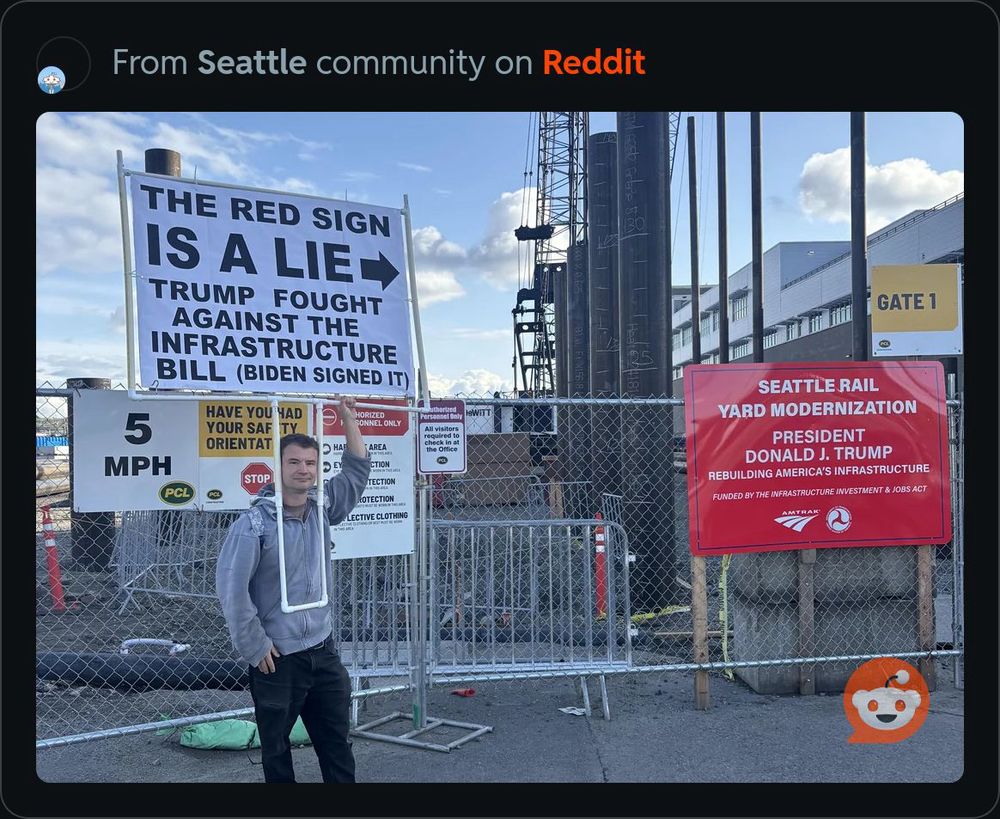 Red sign posted on construction site reads, “Seattle Rail Yard Modernization. President Donald J. Trump. Rebuilding America’s Infrastructure. Funded by the Infrastructure Investment & Jobs Act.” Red sign also features logos for Amtrak and the U.S. Dept. of Transportation.

To the left of the red sign is a man wearing a large white sign that reads, “THE RED SIGN IS A LIE. ➡️ Trump fight against the infrastructure bill (Biden signed it).”