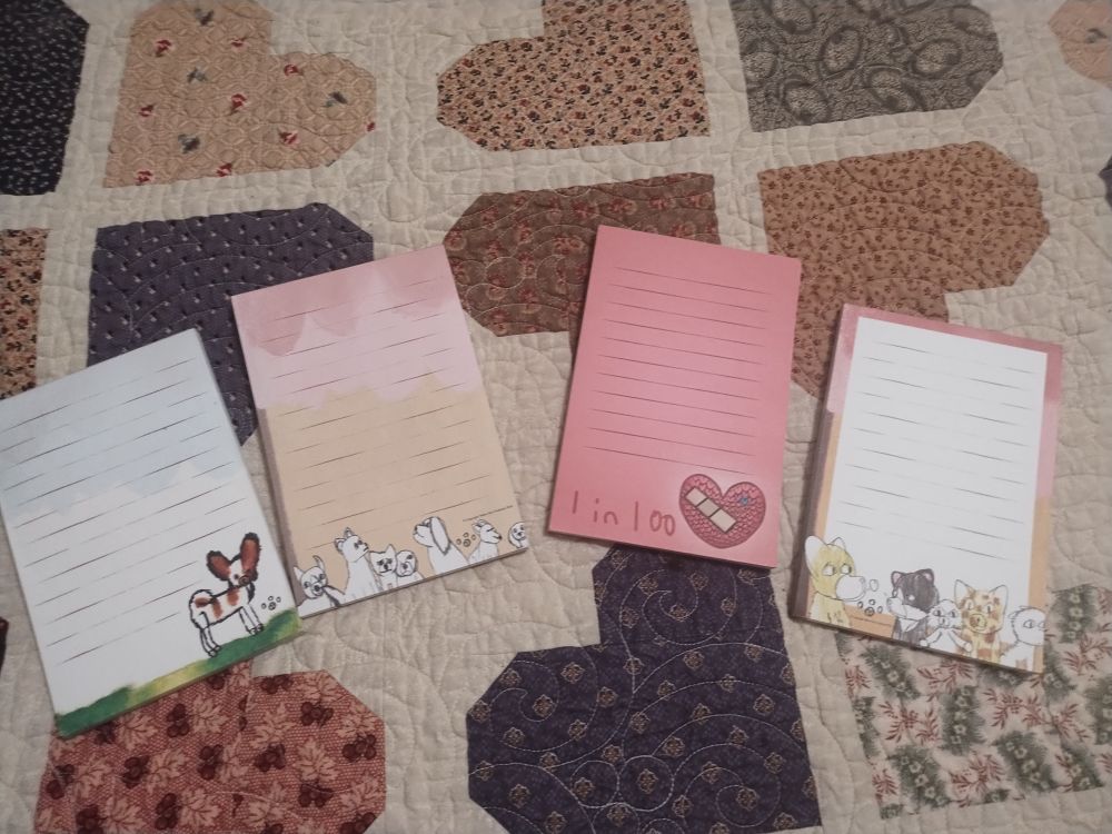 4 notepads laid across on a quilt 
The first one has a dog in the corner.
the 2nd has doubles of dogs on the bottom.
 the 3rd has a heart with a bandage on the bottom right corner with the words "1 in 100" (this won't be one that's available) 
the last has a pink and yellow border with drawings of dogs and cats on the bottom.