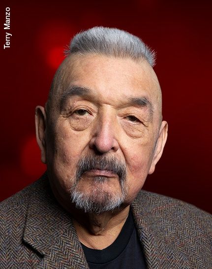 A portrait photo of Graham Greene wearing a tweed blazer sitting in front of a red background taken by the Governor General's Performing Arts Awards for the Lifetime Artistic Achievement Award he was given in 2025.