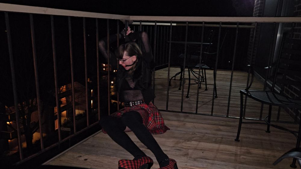 Kira, wearing a black cropped jacket, a sheer spiderweb pattern top over a black bra, a red plaid skirt, black thigh socks, and red plaid high heels. She is gagged with a red ballgag with a black strap. She sitting down, and is handcuffed to the railing of a balcony