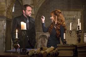 Mark Sheppard and Ruth Connell in Supernatural. Mark in a black suit and Ruth in a green dress. Both standing in a room with lit candles. 