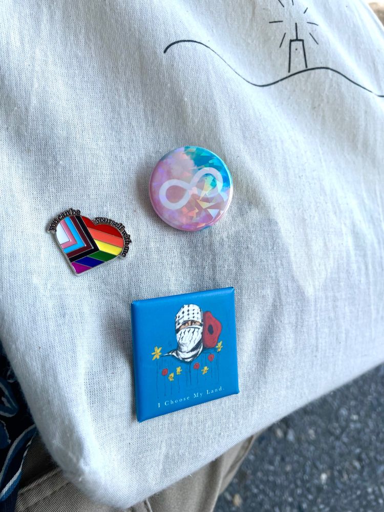 白いトートバッグにニューロダイバーシティプライドと、プログレスプライドと、パレスチナ解放の缶バッジ