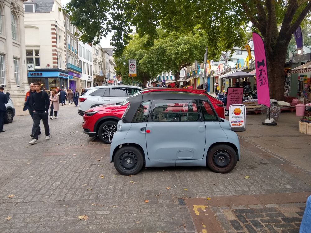 A small grey coloured car parked in a city centre location.