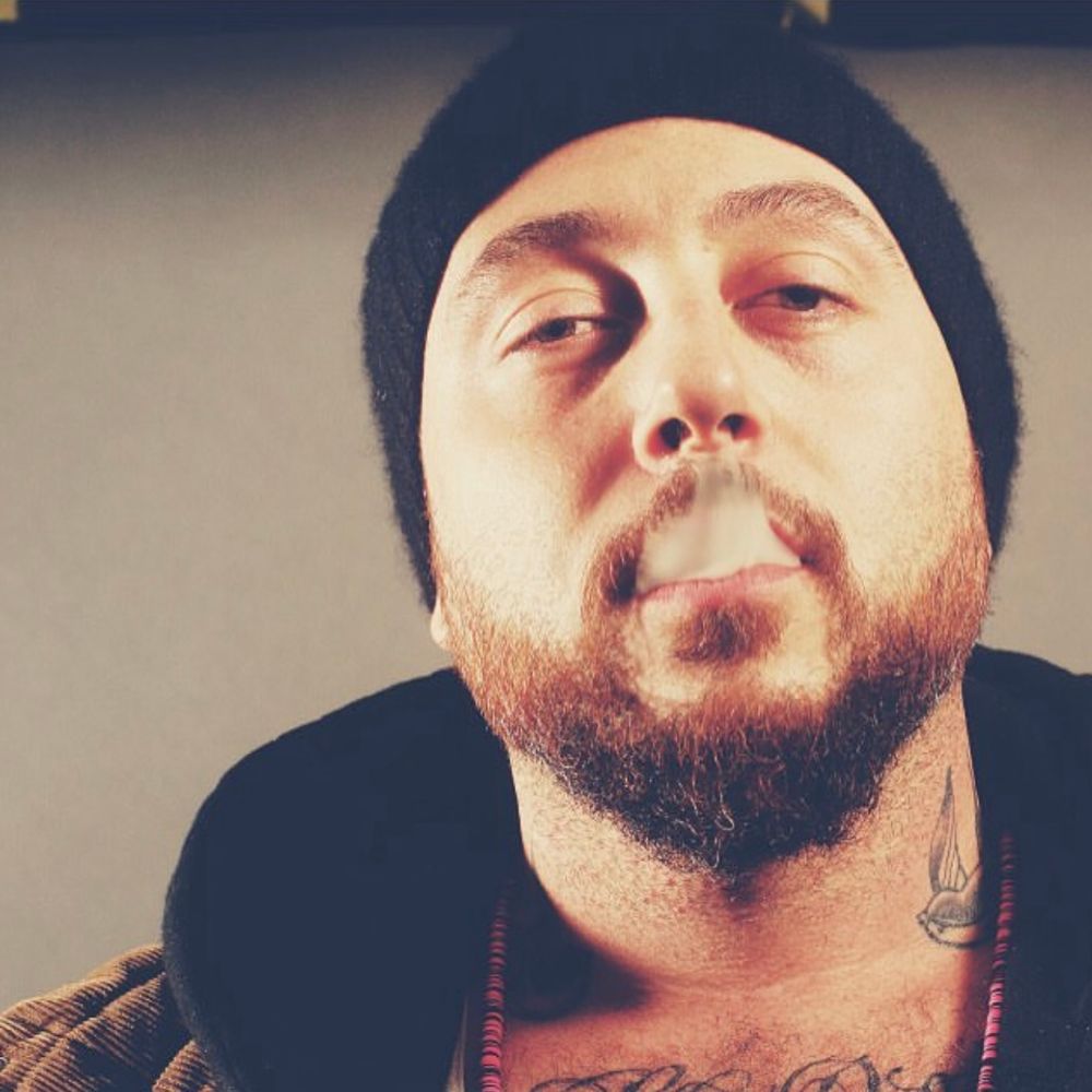 An Hispanic man in his 30s with a medium-sized beard, black skullcap, open hoodie, black-and-red Asian beaded necklace and two neck tattoos blowing smoke from his mouth into his nose 