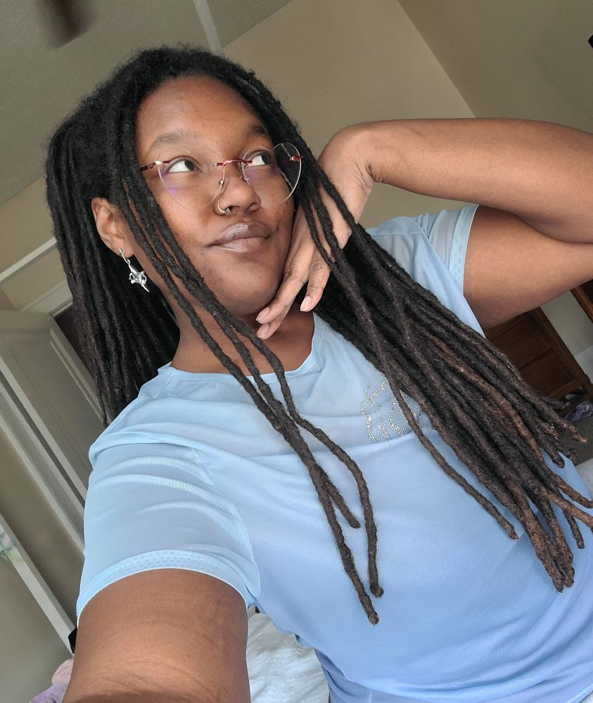Pictured is a species of Bluu, a Black nonbinary baddie. Bluu is wearing a light blue shirt, silver star earrings, and red heart shaped glasses. Bluu has their hand under their chin/cheek and is looking upwards with a smile. They have long locs ending at their stomach, is a fat being with a round face, and warm brown skin. 