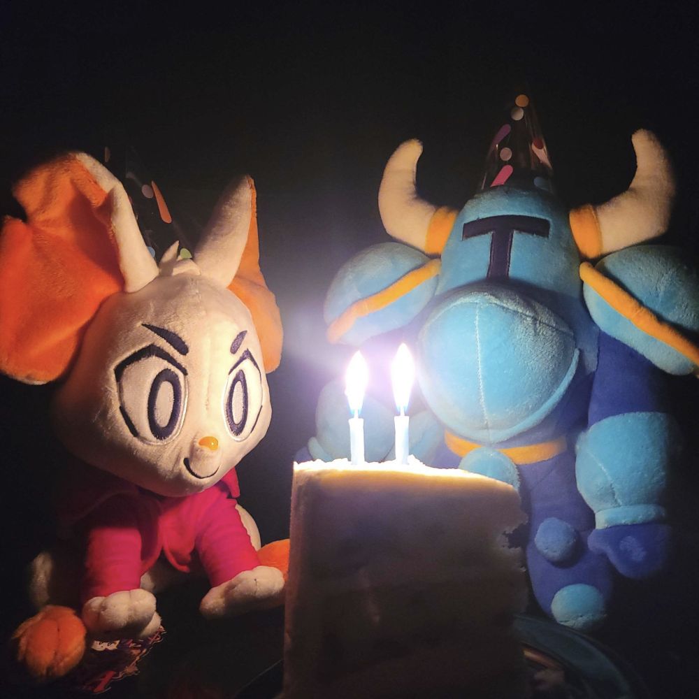A photo of a Mina the Hollower and Shovel Knight plushies sitting in front of a birthday cake with two lit candles. They're both wearing birthday hats. Mina is looking intently at the cake.