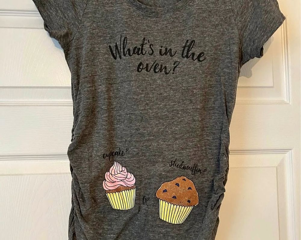 Grey t-shirt with an illustration of a cupcake and a chocolate muffin. Text  on the shirt reads: "What's in the oven? Cupcake or stud muffin?"