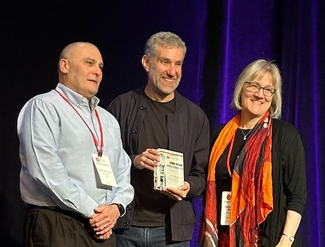 James Fraser, with graying hair and short beard wearing two black shirts and jeans, in front of a curtain holding the award. He is flanked by a man with very little hair wearing a light blue dress shirt and dress pants and and a woman with shoulder-length blond hair and glasses wearing a black top and predominantly red and orange multi-colored and patterned scarf.