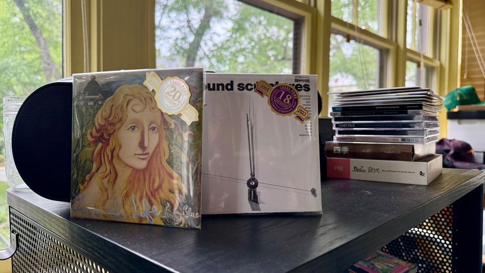 Two CDs from the reissue/bootleg label Creel Pone perched in front of a CD boombox, a stack of CDs behind 