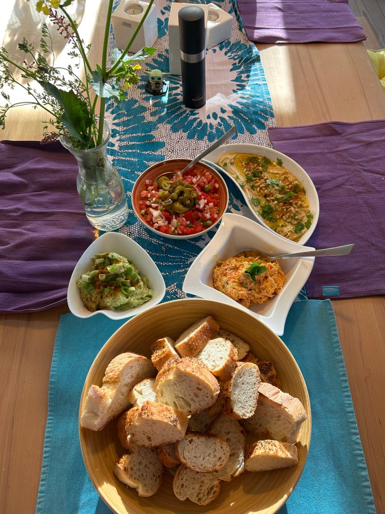 4 Schalen mit Mezze (1x Guacamole, 1x Salsa, 1x Havuc Salatasi, 1x Fasulya Hummus). 1 Korb Brot. Alles steht auf einem Tisch angerichtet, daneben eine Vase mit Feldblumen.