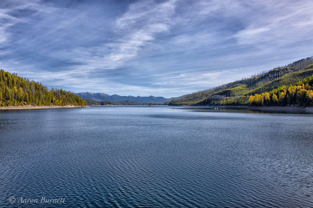 Tranquil lake surrounded by lush green and autumn-colored trees, with mountains and a calm, blue sky in the distance.