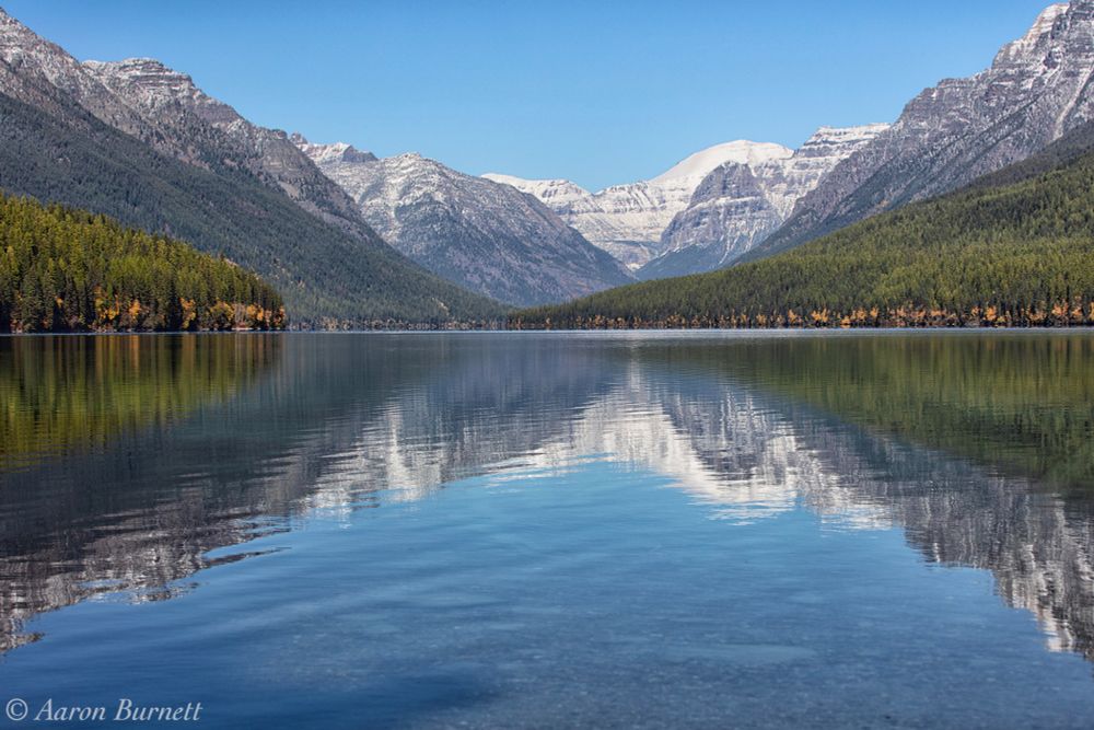 A serene lake reflecting majestic snow-capped mountains and lush pine forests under a clear blue sky.