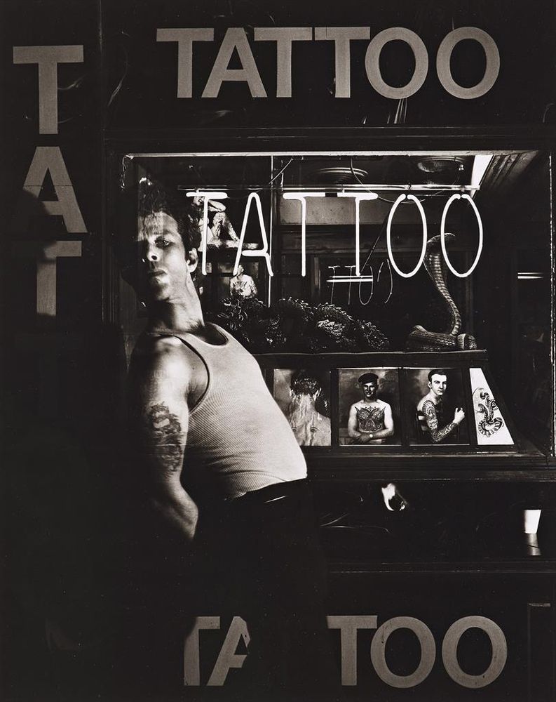 Foto a Tom Waits in piano americano, di profilo, in un negozio di tattoo. Indossa pantaloni e canotta, e si piega all'indietro.
