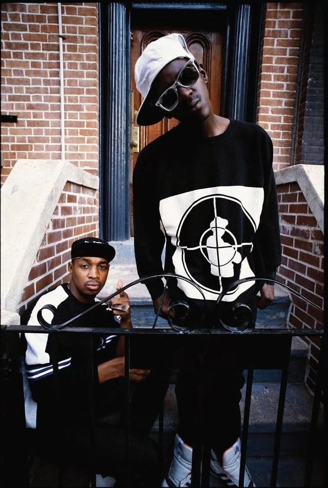 Foto ai Public Enemy sui gradini dell'ufficio della Def Jam, la loro casa discografica.  Chuck D è seduto, con indosso una felpa nera con bordi bianchi sulle braccia. Flavor Flav è in piedi con occhiali da sole, capello da baseball di lato, testa inclinata e indossa una felpa nera con il loro logo.