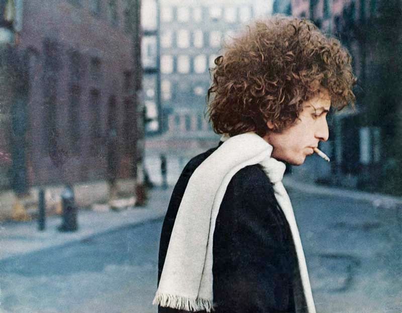 Foto di profilo, a mezzobusto, a Bob Dylan che fuma una sigaretta in una strada di Manhattan. Ha un cappotto nero e una sciarpa bianca al collo.