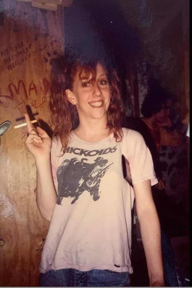 Foto a mezzafigura a Teresa Nervosa in un locale con muri tutti scritti, con una sigaretta in mano, un sorriso da foto, e una maglietta degli Hickoids