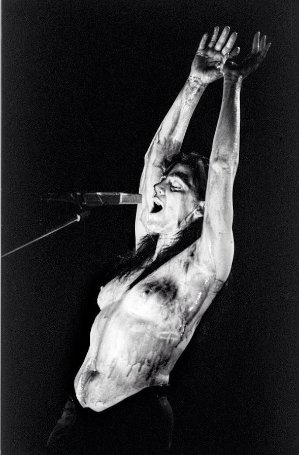 Diamanda Galas sul palco, in topless, con le mani alto e con il corpo ricoperto da qualche sostanza colante, mentre canta