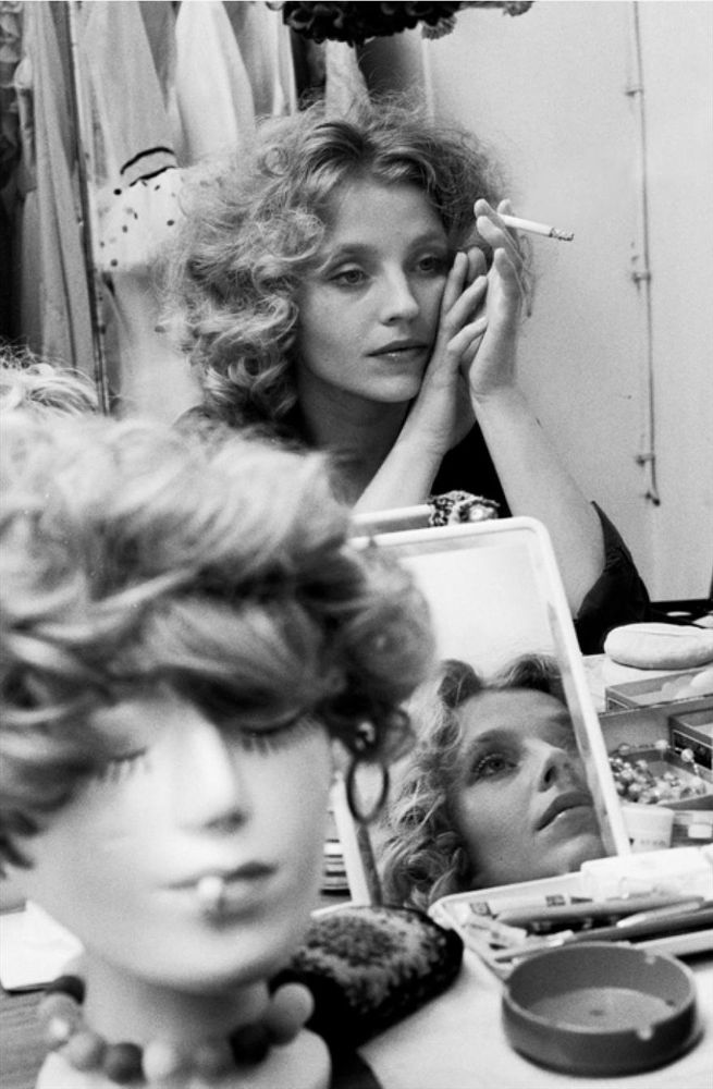 Foto a Hanna Schygulla nel camerino davanti allo specchio. Ha un'espressione riflessiva e rilassata mentre tiene una sigaretta nella mano destra e appoggia il mento sulla sinistra. In primo piano, è visibile una testa di manichino con una parrucca riccia e una collana di perline, anch'essa con sigaretta in bocca. La toeletta è ingombra di vari oggetti, tra cui uno specchio in cui si riflette il volto della donna, un posacenere e contenitori che sembrano contenere trucchi o piccoli accessori. 