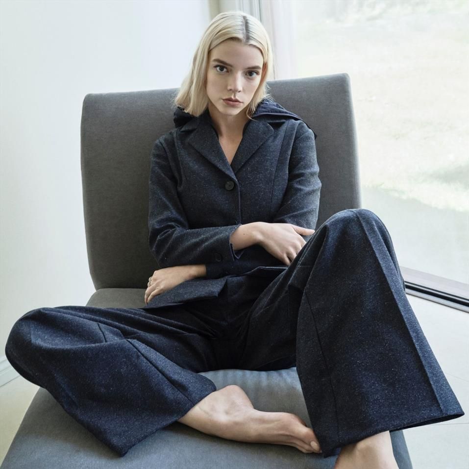Anya Taylor-Joy seduta su una poltrona moderna grigia vicino a una grande finestra, indossa un completo blu scuro con giacca e pantaloni larghi. È a piedi nudi e guarda dritto in camera con espressione seria, trasmettendo un’atmosfera di calma e introspezione.