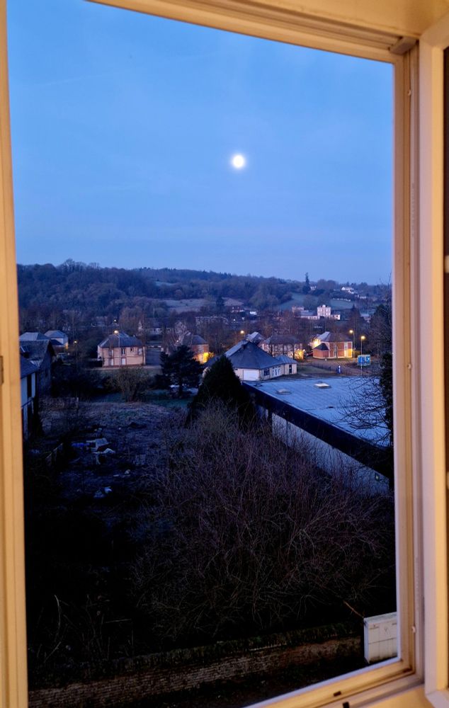 Vue par la fenêtre du 2e étage au petit matin, la pleine lune dans le ciel bleu nuit, les toits des maisons et les lumières du village