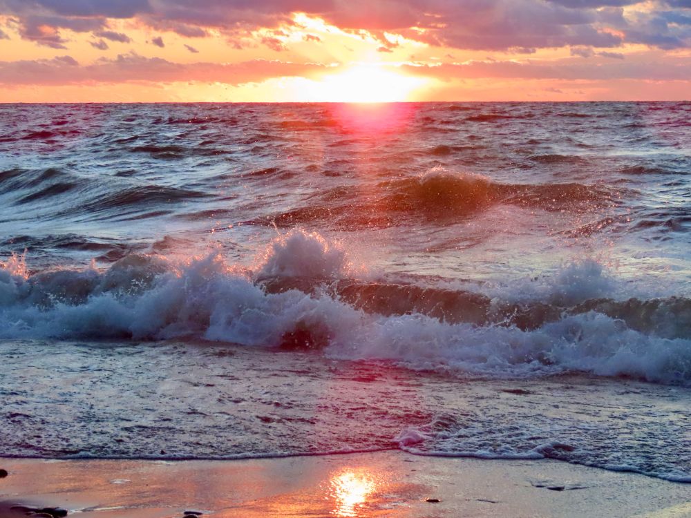 Photo of the sun setting at the horizon along Lake Erie. Moderate waves are crashing along the beach. The sky is shades of pinks and yellows and golds that are reflected in the water. 