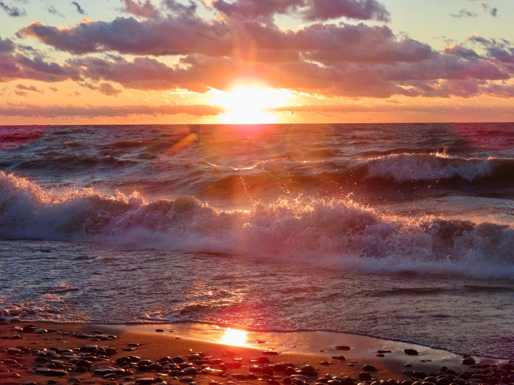 Photo. Sunset over Lake Erie. The sun is on the  horizon, shining in bright golds while the sky and lake reflect reds and oranges. There’s wet sand in the foreground with large beach pebbles, also reflecting the colors of the sky. A row of gray and purple clouds in the sky mimic the shapes of the waves below. 