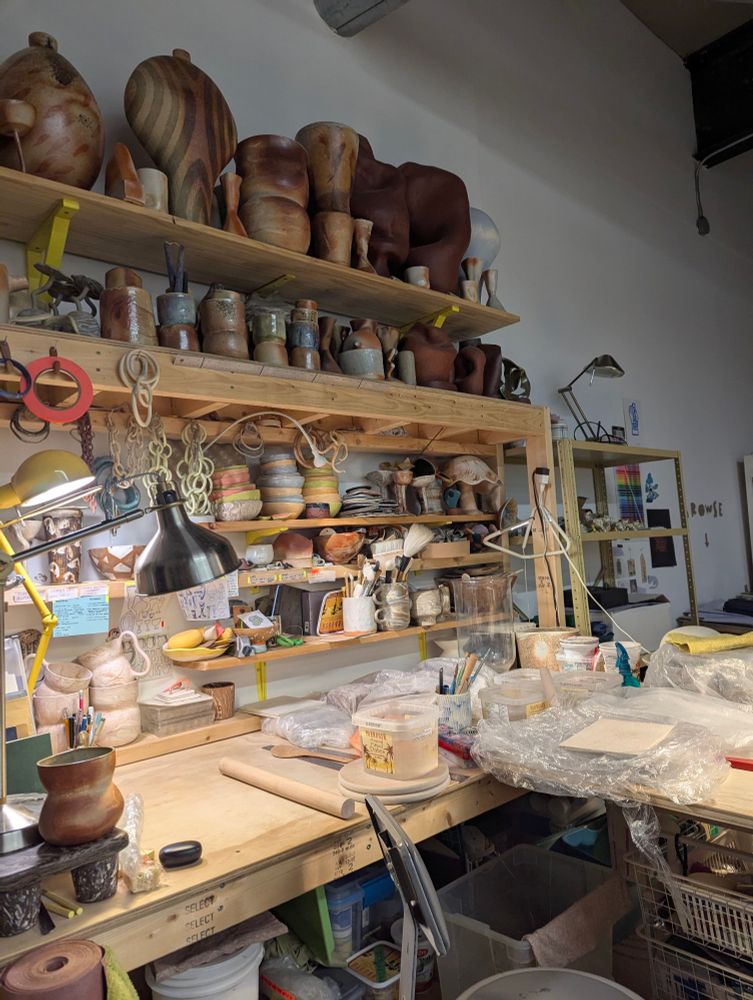 Work desk with shelves and stacks of ceramic bowls, cups, sculptures and miscellaneous clay objects. The desk surface has wrapped up works in progress.