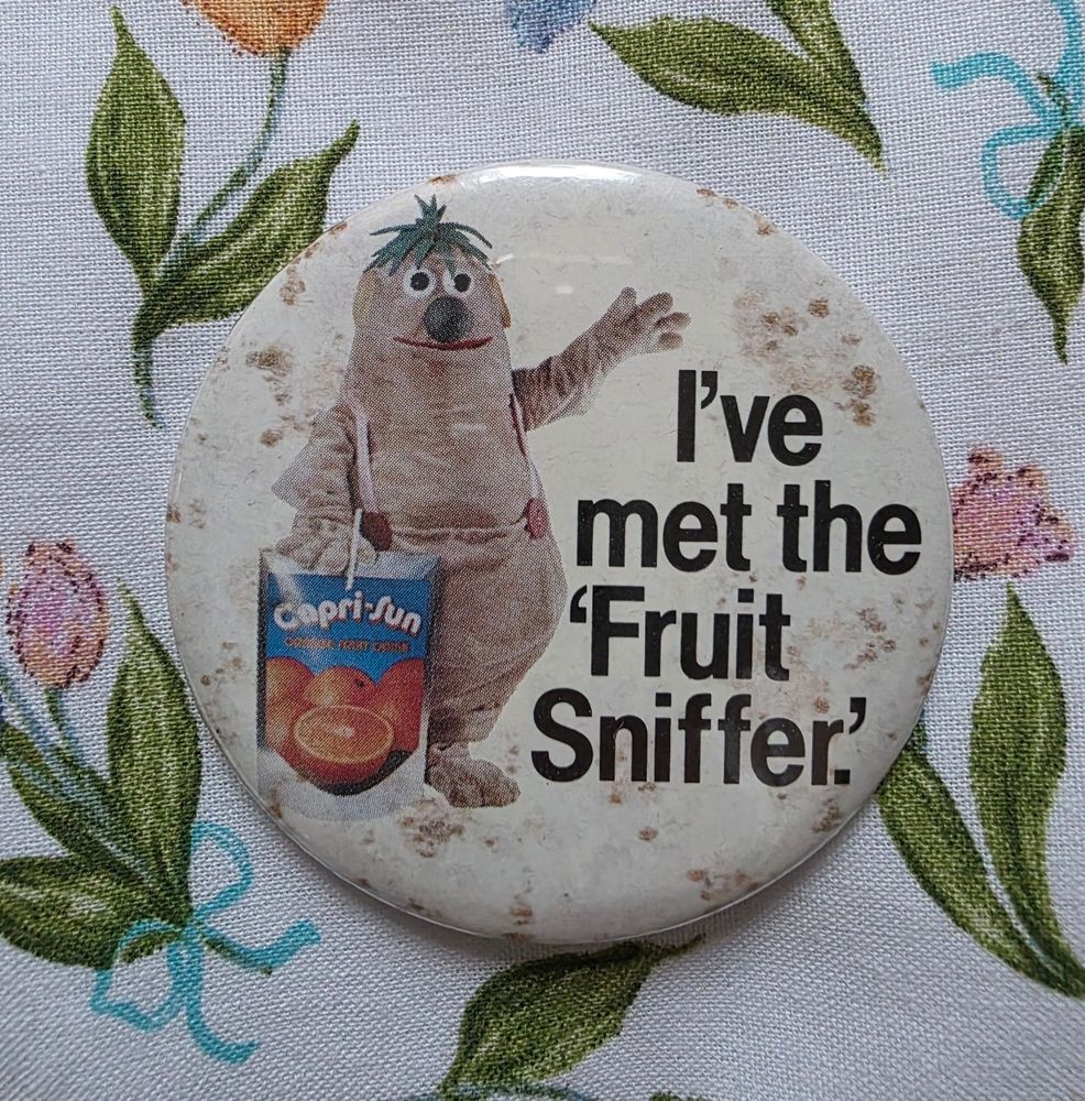 A round, slightly stained promotional button featuring a costumed character holding a large Capri-Sun juice pouch. The character is beige, with a round body, green hair-like tufts, and a big red nose. Bold black text next to the character reads: "I've met the 'Fruit Sniffer.'" The button is placed on a floral-patterned fabric background.