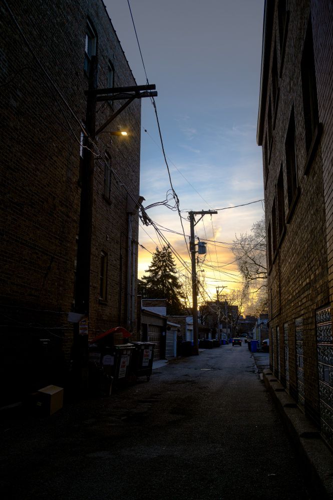 Looking down a dark ally, the sun sets over distant houses.