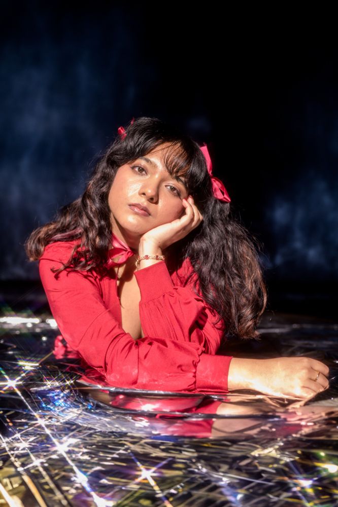 A young woman lying down on a sheet of shining silver rests her head in her hand as she looks into the camera. 