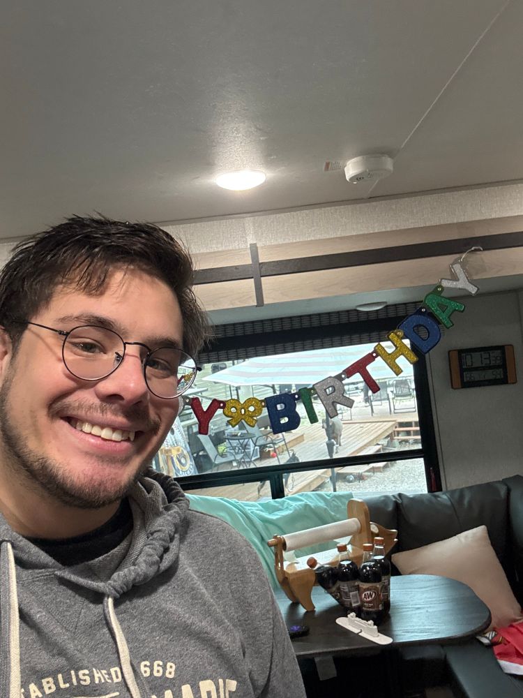 Young man with glasses and short hair standing in front of a window and table, with a “Happy Birthday” banner behind him. 