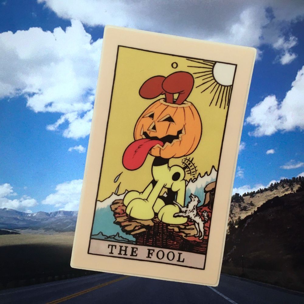 A picture of a sticker on a landscape background. It is The Fool tarot card, but the fool is Odie wearing a jack'o'lantern head.