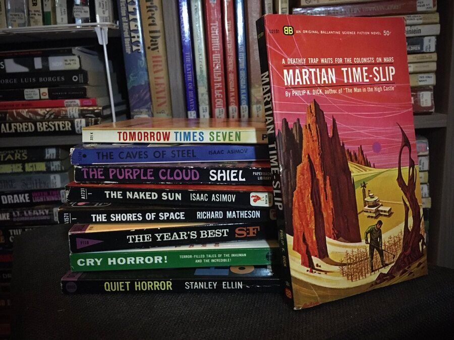 A stack of mid-century mass-marked paperback books: Tomorrow Times Seven, The Caves of Steel, The Purple Cloud, The Naked Sun, The Shores of Space, The Year's Best SF, Cry Horror!, Quiet Horror. Leaning against the stack is Martian Time-Slip by Phillip K. Dick, the cover showing a man hoeing a crop in an alien landscape, a futuristic looking house in the valley below him.
