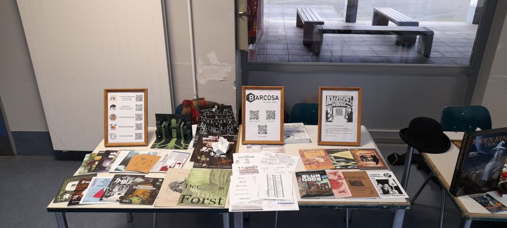 Ein Foto vom Stand, der aus 2 kleinen Tischen voll mit Fanzines besteht und ein paar kleinen Aufstellern mit Scancodes
