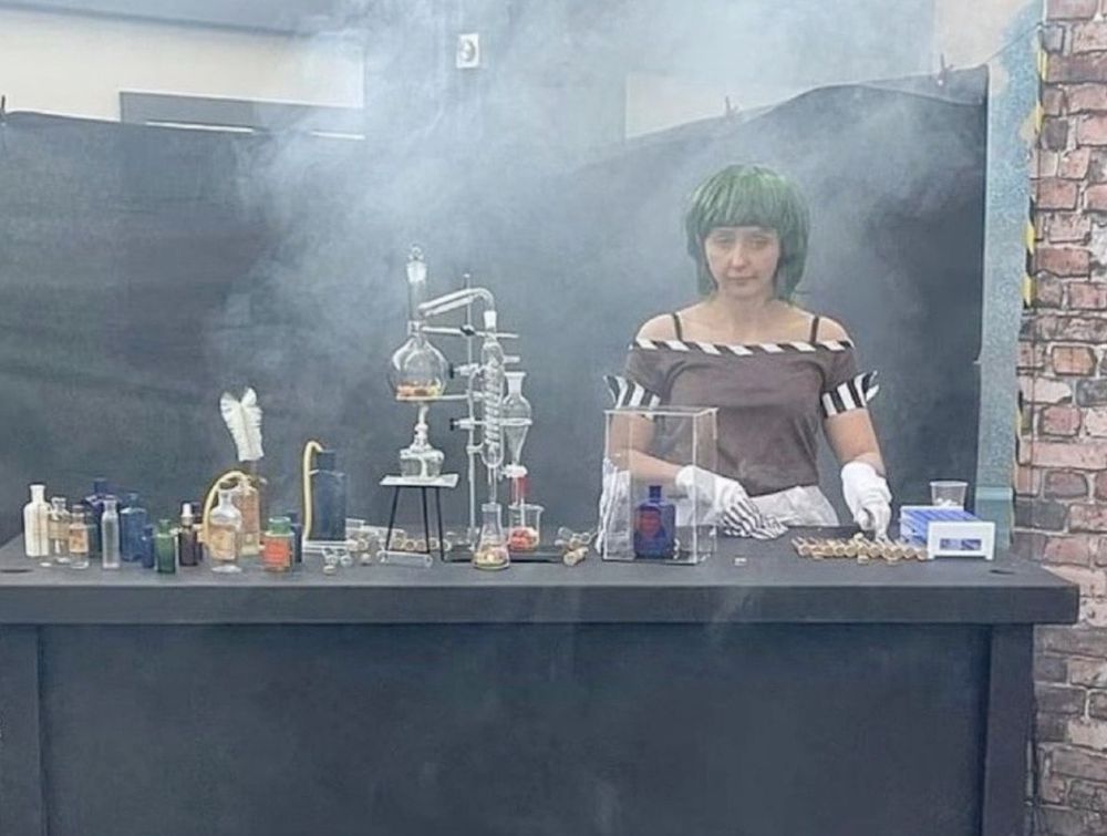 a photo from the viral "Willy's Chocolate Experience" event that was a notorious failure: the photo is of a woman in a green wig standing in front of a poorly-hung black sheet. She has a table with haphazard "lab equipment". There is a fog machine seemingly pointed at her. Her expression is exhausted. The overall effect is both underwhelming and embarassing
