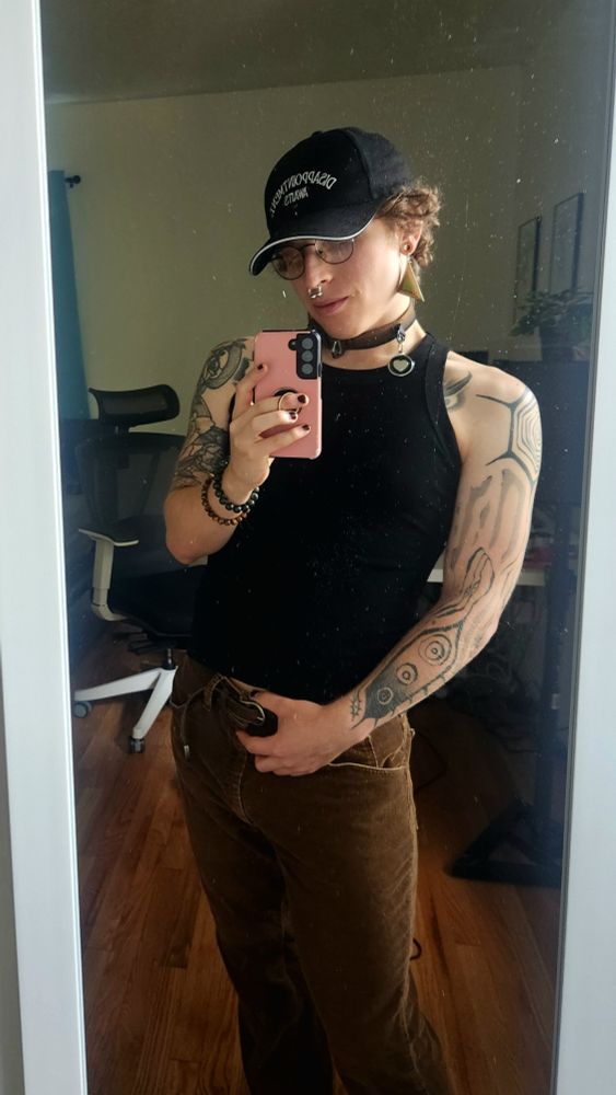 Ina, a trans femme, dressed in brown corduroy pants and a tight black tank top
