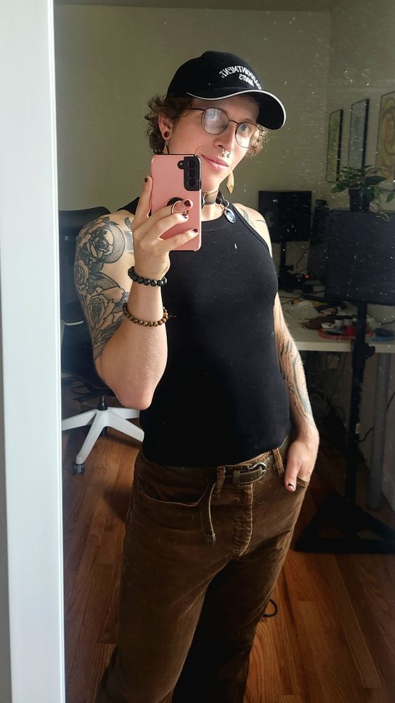 Ina, a trans femme, dressed in brown corduroy pants and a tight black tank top