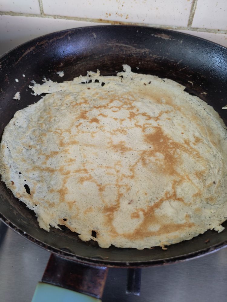 Somewhat untidy crepe-style pancake in a frying pan, turned onto the second side so the darker pattern is visible from cooking 