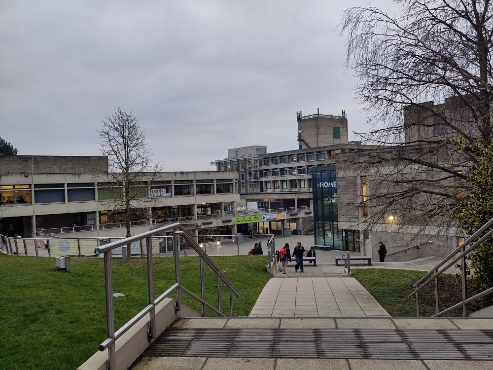 Looking across the UEA square from the top of the steps. Grey concrete and grey sky in all its brutalist splendour and a splash of colour from green grass and a student's red bag