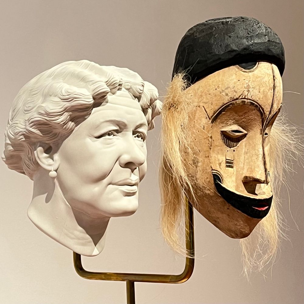 Eine Skulptur aus Gips zeigt eine ältere Frau mit europäischer Flechtfrisur mit Perlenohrringen. Davor ist mit Abstand eine Maske aus Zentralafrika montiert. Beide Darstellungen der Gesichter sind idealisiert. (fehlerfrei). Je nach Standort sieht man nur die Maske von vorne über beide seitllich bis Kopf von hinten (Blick auf den Haarknoten)