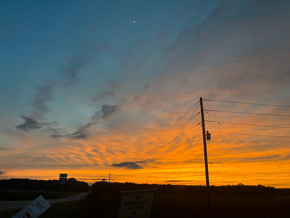 Rich orange striated clouds against darkening blue sky,  sunset over an open field at a truck stop in a rural part of northern North Carolina, upper center of pic has the moon and Venus, I think, shining in distance.