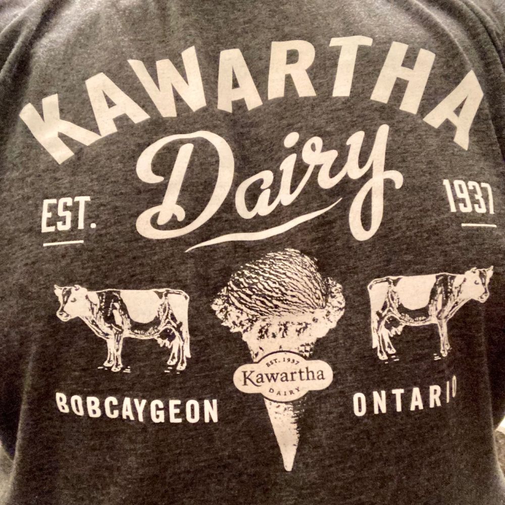 A grey t-shirt with “Kawartha Dairy Est. 1937 Bobcaygeon Ontario” in white around a picture of an ice cream cone and two cows.