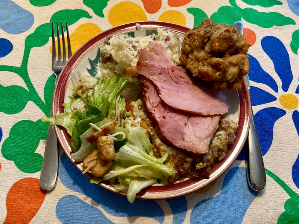 A plate of Thanksgiving dinner. The plate is white stoneware with a green holly print and red border. The food includes, clockwise from top, mashed potatoes, two slices of ham, stuffing (in the form of a muffin), green-bean casserole under the ham, and a tossed iceberg-lettuce salad with croutons. The entire plate is on a floral-print tablecloth.