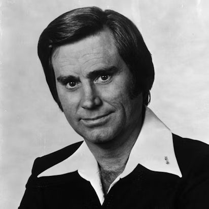 Country Music Hall of Famer George Jones, probably from the 1970s. The picture is a black-and-white head shot of Jones wearing a dark suit or sport jacket over a white shirt with a wide, open collar in front of a light background. Jones looks only mildly psychotic and/or high in the photo.