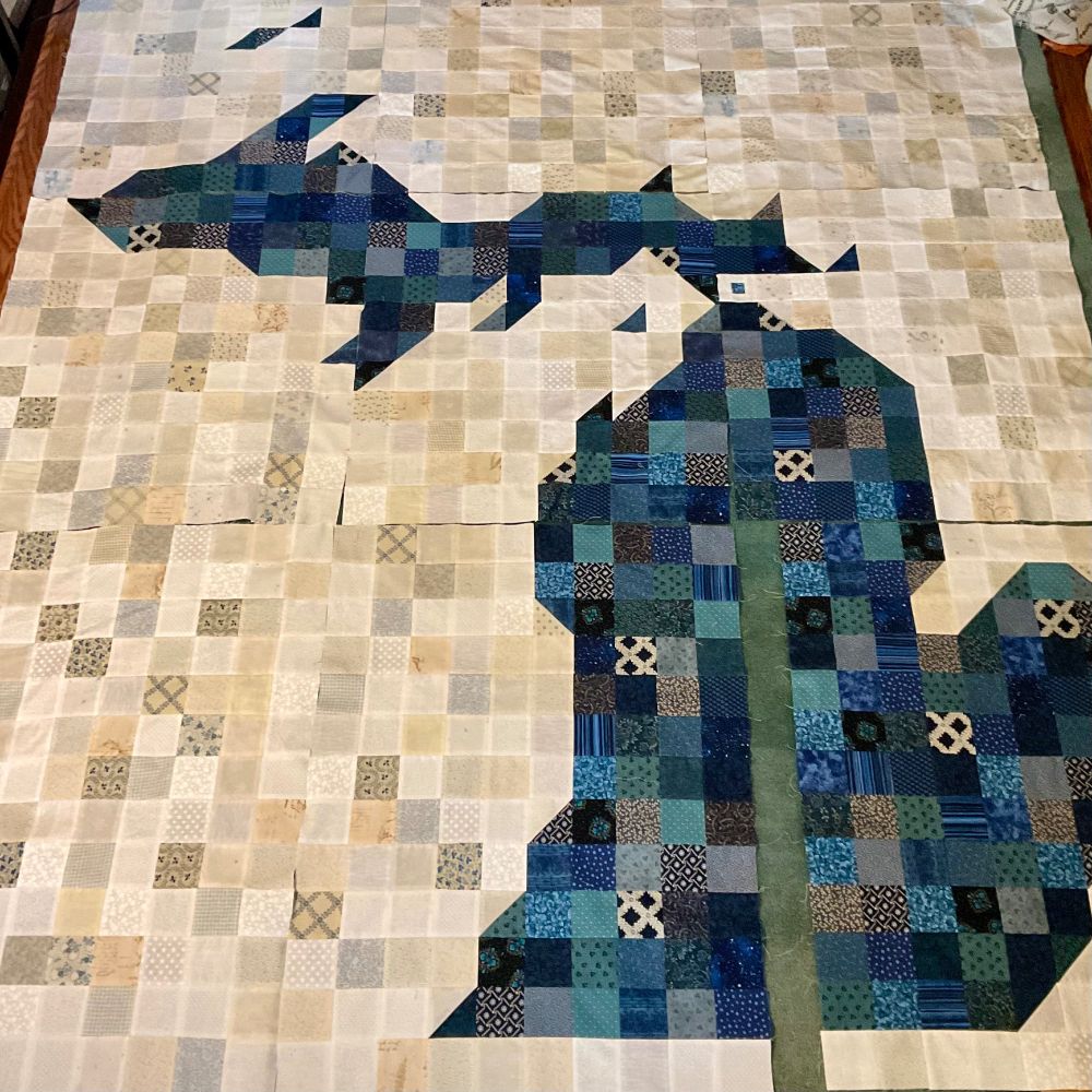 A partially completed quilt topper comprised primarily of 3” squares. The state of Michigan is rendered in a mix of blue patterns while the background is in neutrals. A strip of 13 squares through the heart of the lower peninsula is missing.