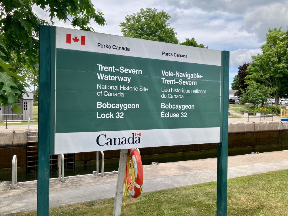 A Parks Canada/Parcs Canada sign reading, 
“Trent-Severn Waterway/Voie-Navigable-Trent-Severn
“National Historic Site of Canada/Lieu historique national du Canada
“Bobcaygeon Lock 32/Bobcaygeon Écluse 32”

The sign is green with white text. Parks Canada/Parcs Canada at the top and Canada at the bottom are in black text on white bands. The sign is located on a grassy area in front of the canal. Trees, parkland, and vehicles can be seen on the other side of the canal.