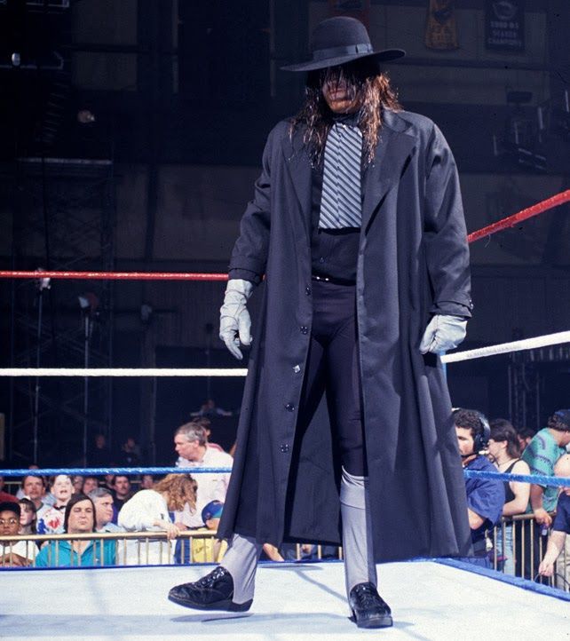 WWF wrestler Brian Lee standing in-ring in character as the fake Undertaker. He is dressed as a stylized undertaker with black pants and shirt, a long black coat, and a black wide-brimmed hat hiding most of his face. A 1990s wrestling crowd is visible outside the ring.
