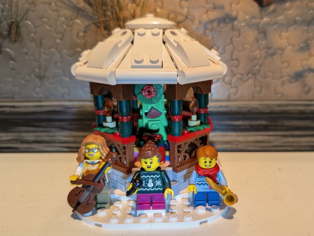 A close-up photo focuses on a LEGO winter scene displayed on a mantle, featuring a brown and white gazebo with a Christmas tree inside. Three minifigure musicians stand in the snow as a silent holiday band. On the left, a figure with long hair and glasses plays a cello. In the center, a singer wearing a green sweater with a snowman pattern holds a microphone. On the right, a musician in a blue sweater and red scarf plays a gold saxophone. The background shows the texture of a jigsaw puzzle on the wall behind the display.