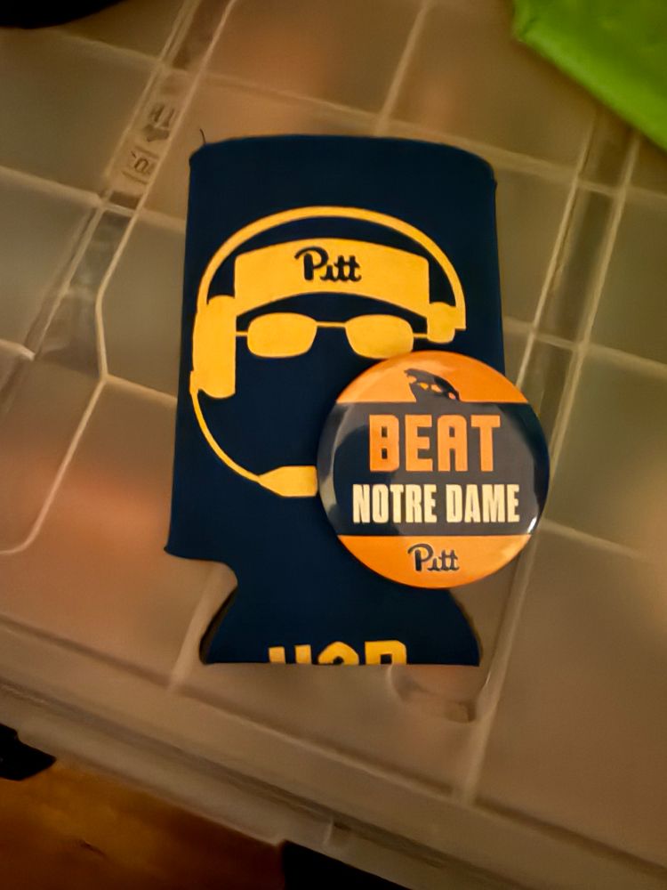 A beat Notre Dame pin on top of a koozie with a silhouette of Pat Narduzzi’s glasses, visor, and headset